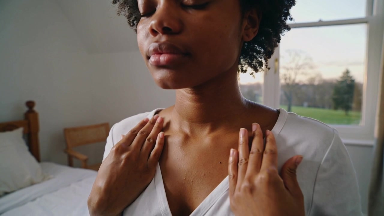 Les mains sur la clavicule, une femme ajuste son haut à col en V dans une chambre près d'une fenêtre, laissant apparaître une bretelle noire