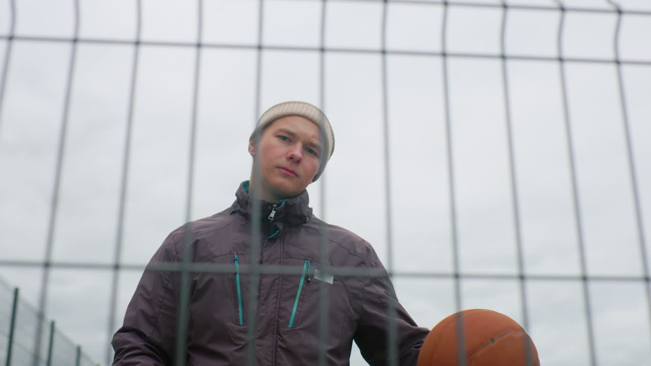 young white male with basketball behind fence, standing on outdoor court in beanie and jacket, holding ball with confident yet pensive look, overcast sky, metal grid foreground, urban streetball vibe