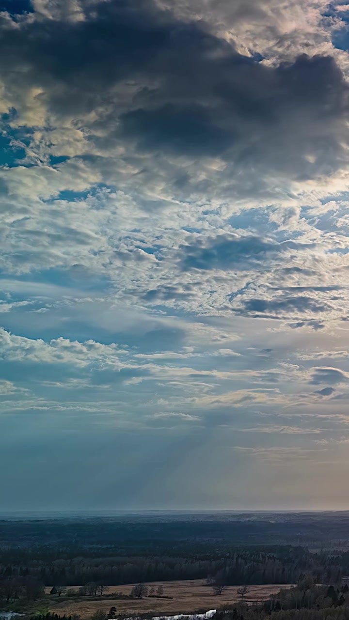 Dramatic layered clouds spreading across blue sky above distant rural terrain, aerial vertical static as backlit rays dance