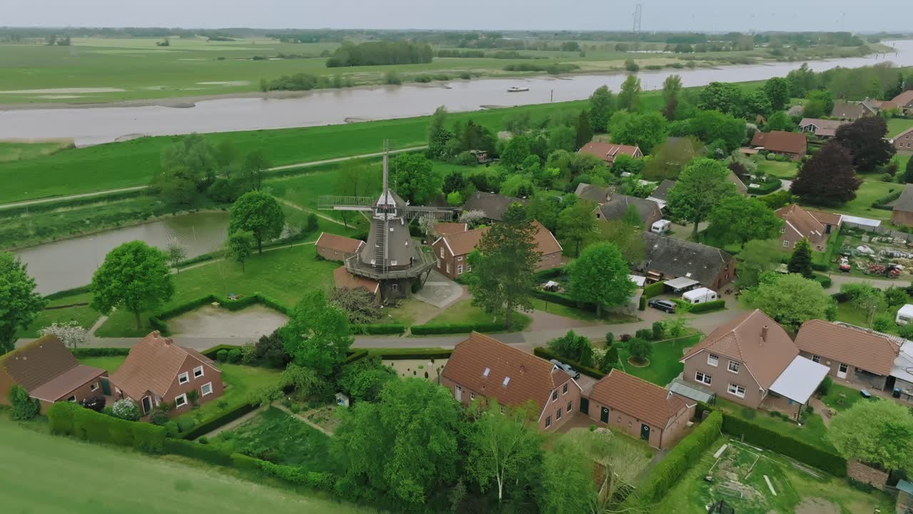Descending drone shot showing green fields, traditional homes and a windmill. Lighting is soft and diffused under cloudy skies.