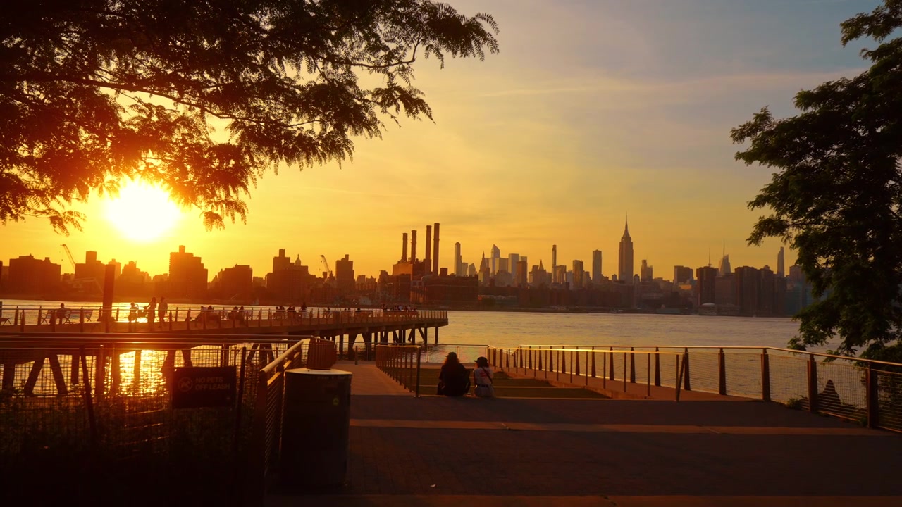 Horizonte de Manhattan através do East River ao pôr do sol. Galhos de árvores emolduram píer, pessoas e reflexos na água.