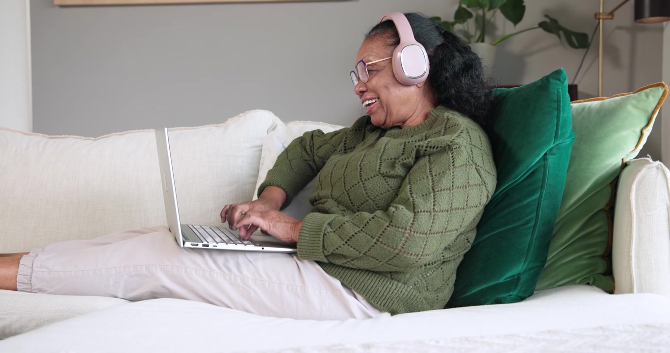 Senior African woman typing on silver laptop on sofa, reaching for pink headphones to hear audio