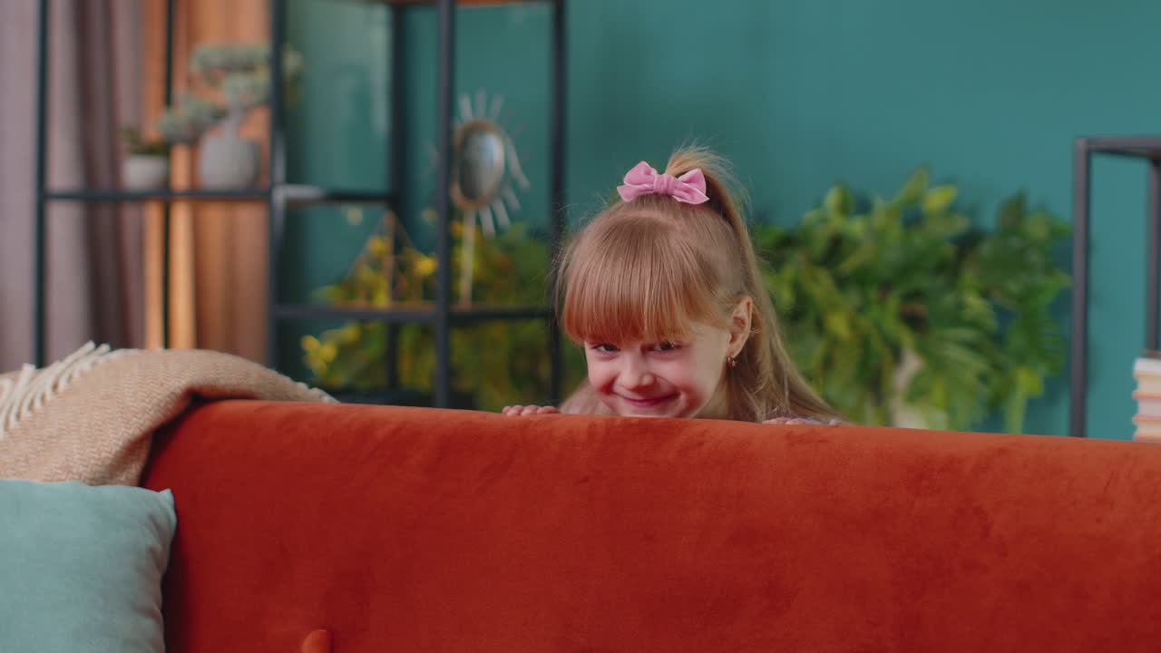 Child girl playing hide and seek peekaboo game near sofa at home alone looking at camera and smiling