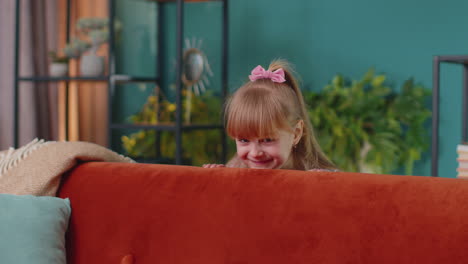 Child-girl-playing-hide-and-seek-peekaboo-game-near-sofa-at-home-alone-looking-at-camera-and-smiling