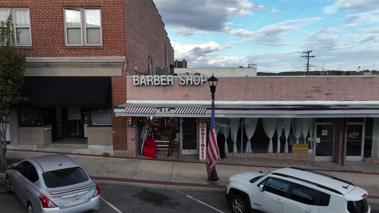 Barber Shop in small american city with hanging flag of USA on lantern. Parking cars along street during autumn day. Aerial orbit shot.