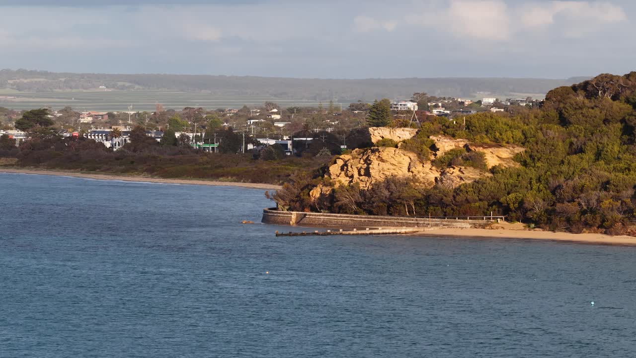 Daytime camera pan reveals rocky headland, calm bay, distant houses, and soft natural lighting