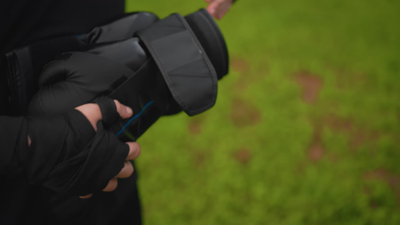 Utendørs votteøvelse med tatovert utøver og trener som holder puter på en gressbane, nærbilde av armtatoveringer og justering av stropper, intenst fokus på grep og målnøyaktighet under padarbeid