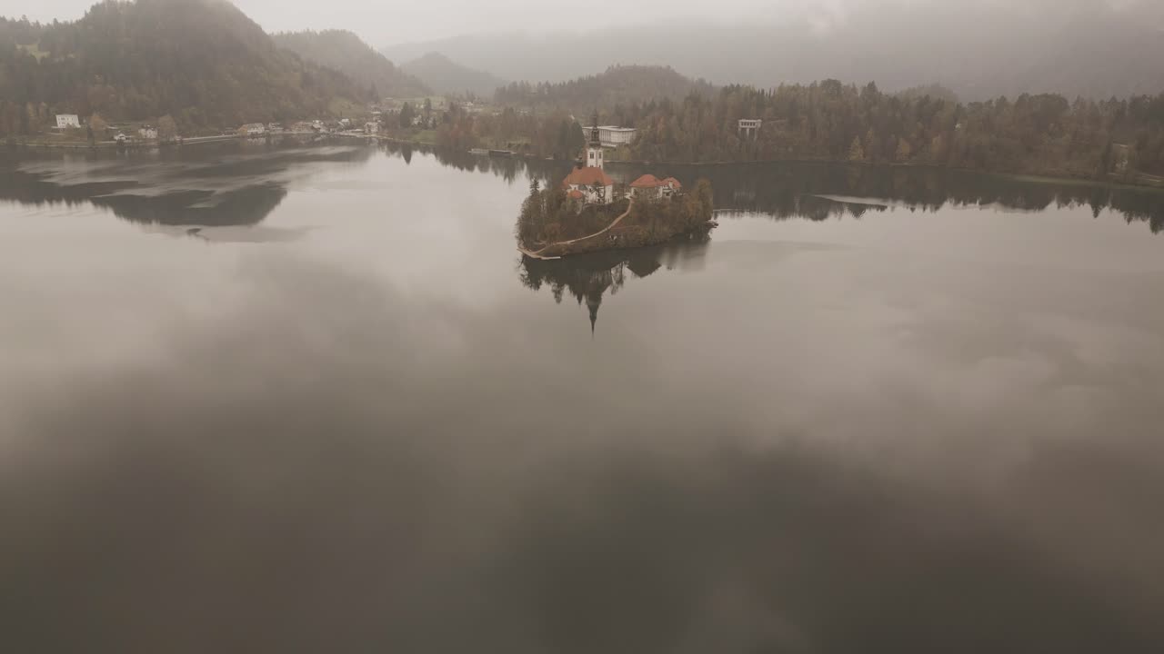 Vista drone sul Lago di Bled, Slovenia, in una giornata autunnale nuvolosa: nebbia leggera si posa sul lago tranquillo, circondato da alberi autunnali colorati, con il Castello di Bled arroccato su una collina e montagne distanti.