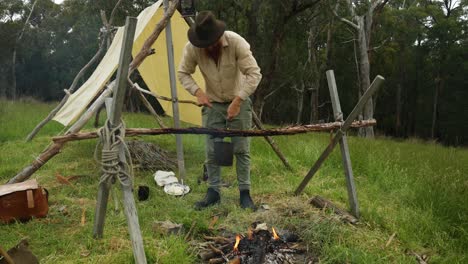 australian bushman hangs up a billy over a campfire in the bush by his historical hut