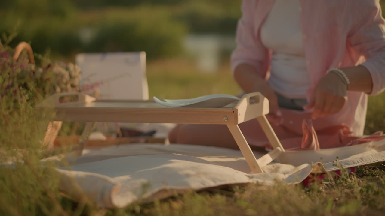 바에 앉아있는 여자가 피크닉 테이블에 접시를 놓고 그녀의 에 인 바구니에 꽃을 놓고, 그녀는 평범한 옷을 입고, 이 많은 풀에서 평화로운 피크닉을 즐기고 있습니다.