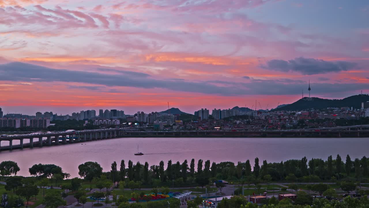 Sweeping aerial view of Banpo Bridge and Han River with Namsan Tower rising above Seoul's skyline, vivid pink and orange sunset clouds glowing over Hangang Park