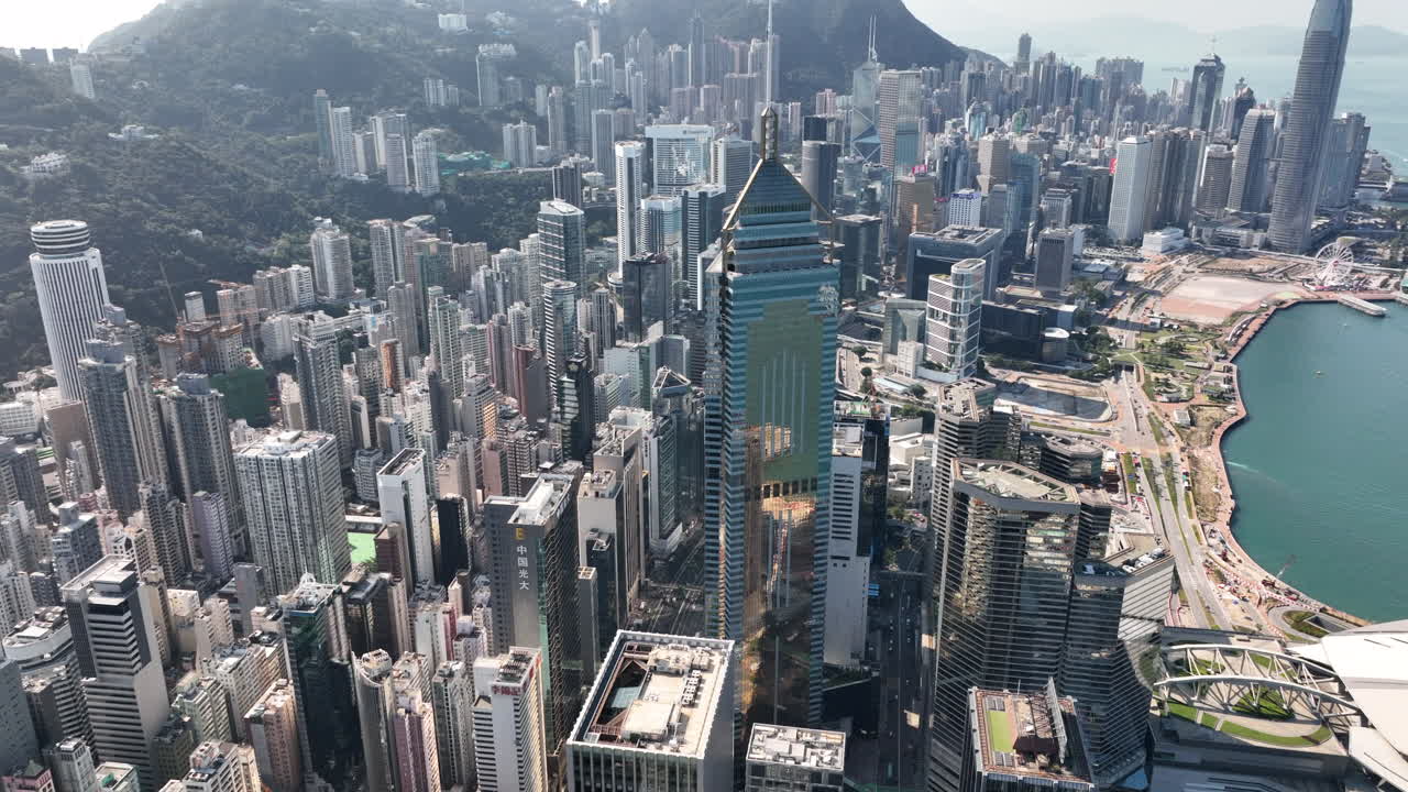 Krouživý letecký záběr za jasného dne na čtvrť Wan Chai s výrazným mrakodrapem Central Plaza v popředí a centrální finanční čtvrtí v pozadí.