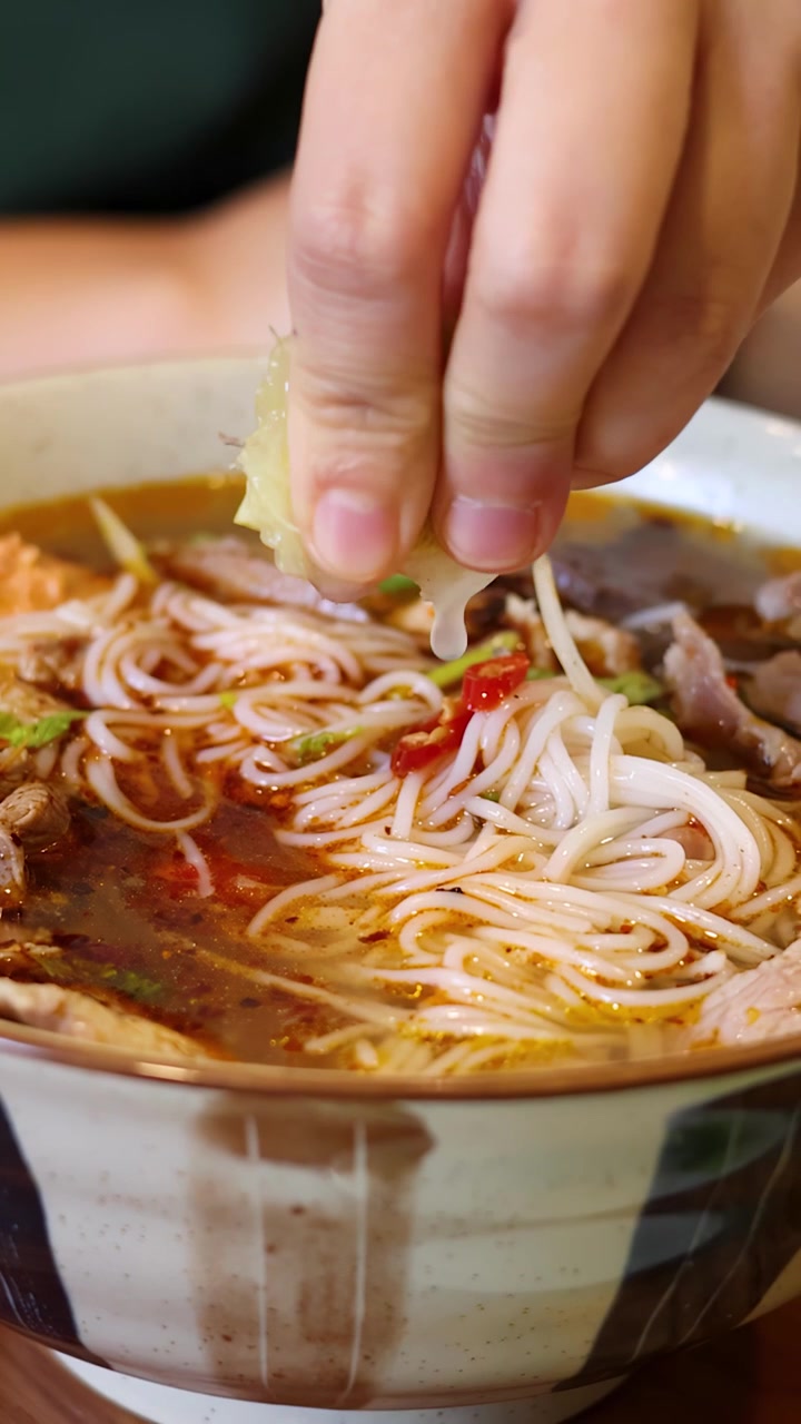 Primo piano di mani che spremono succo di lime su una ciotola fumante di noodles tradizionali con manzo.