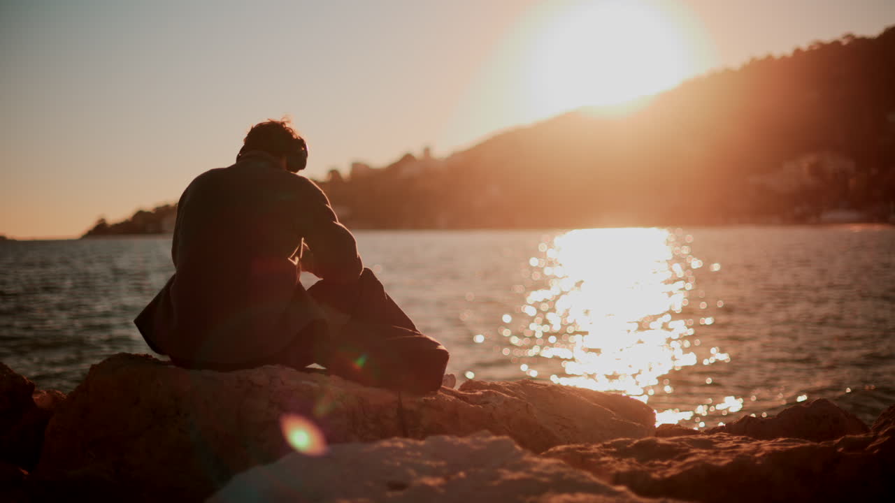 Un uomo seduto da solo su delle rocce costiere, che guarda in basso mentre il sole tramonta sul mare e sulle colline, creando un’atmosfera calda e contemplativa