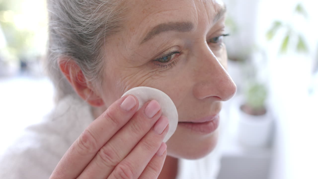 Applying skincare product, mature woman gently cleansing face with cotton pad at home