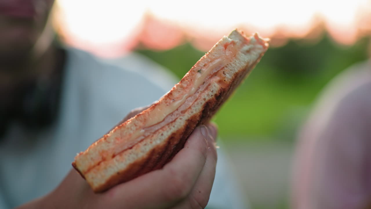 Partial view of person holding sandwich, with another person blurred in the background, outdoor setting with natural lighting, capturing relaxed and casual moments while eating in nature