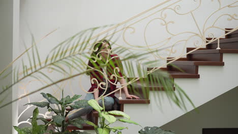 young woman rests on apartment staircase. relaxed female home owner poses for camera sitting on stairs with forged handrails. luxury residence