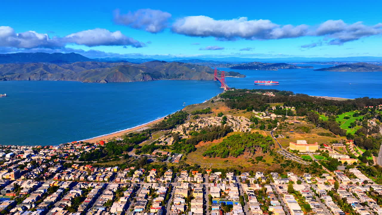 Vista aerea del Golden Gate Bridge che collega l'area di Presidio a San Francisco. Una grande nave portacontainer passa sotto il ponte nelle acque blu della baia. Colline verdi e quartieri urbani.