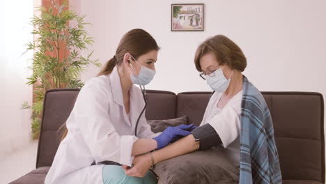 female doctor takes the pulse of an elderly patient sitting on a sofa