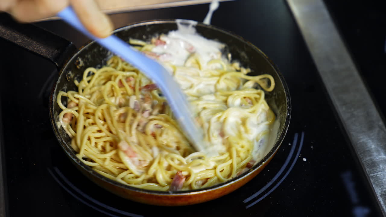Preparation of a delicious dish in frying pan. Cooking and stirring the spaghetti with white sauce in the frying pan. Close-up.
