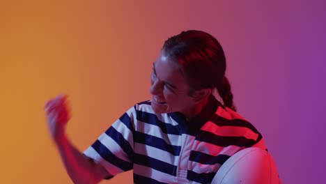 happy caucasian female rugby player raising hands with rugby ball over neon pink lighting