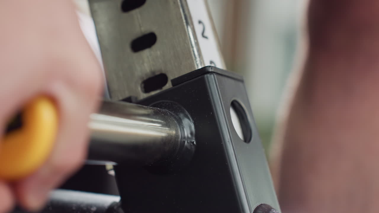 Side view of hand adjusting bench height on weight machine seat, pulling yellow pin to reposition metal lever, showcasing mechanical precision on sleek gym equipment under bright lighting