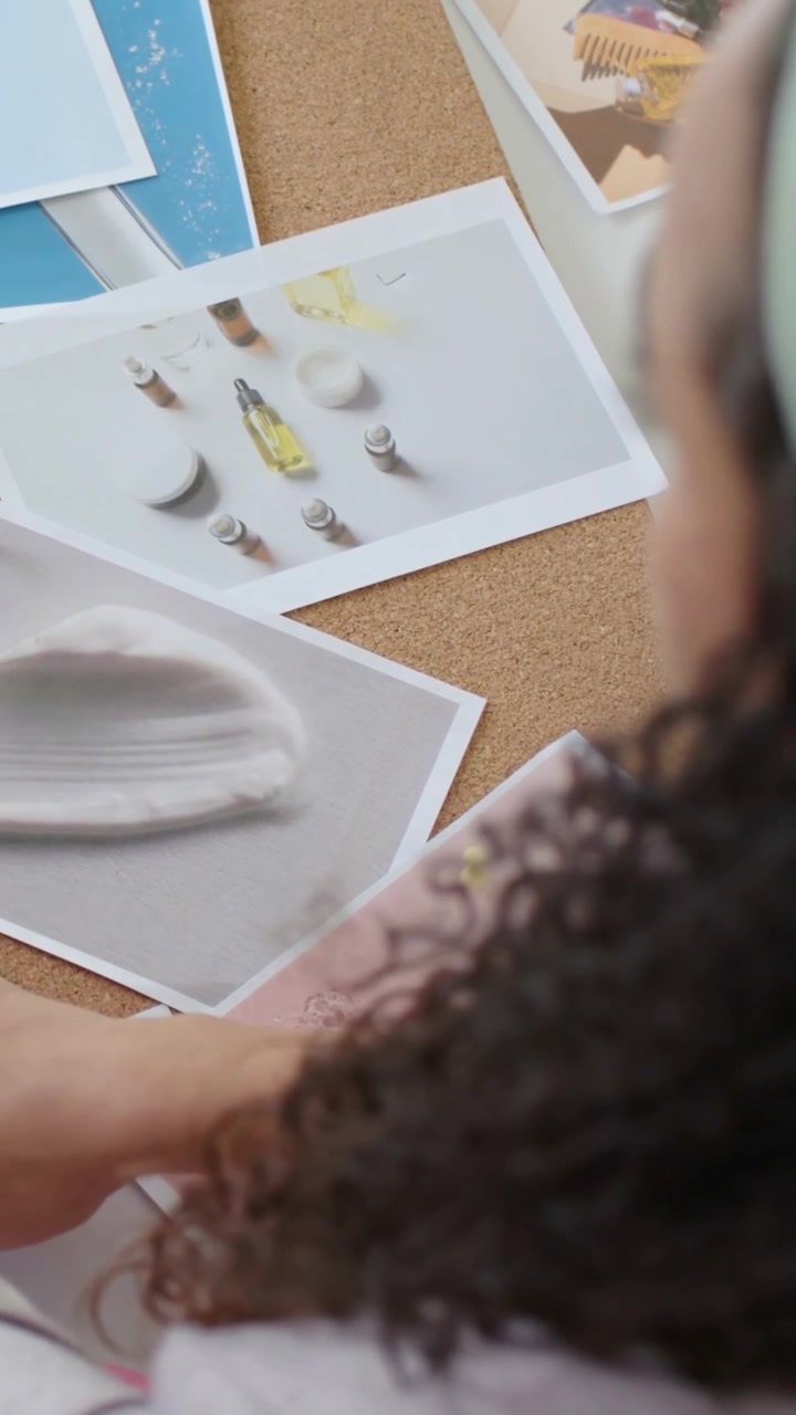 Video verticale: Donna che inizia il layout, appuntando stampe su sughero con spilli, ispezionando il telefono vicino a delle décolleté.