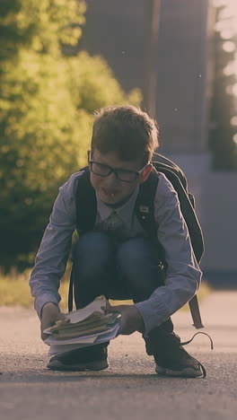 schoolboy wearing glasses with large backpack collects exercise books and sheets on road on sunny day slow motion