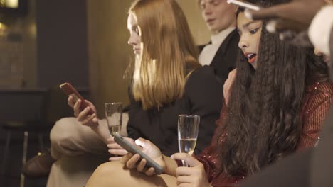 Group of friends dressed in stylish clothes sitting on the sofa while using smartphones and drinking champagne during new year eve party