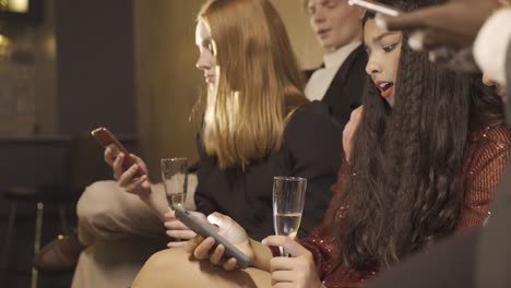 group of friends dressed in stylish clothes sitting on the sofa while using smartphones and drinking champagne during new year eve party