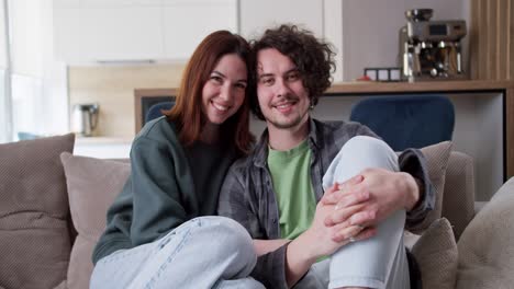 Portrait-of-a-happy-couple-brunette-girl-hugging-her-boyfriend-with-curly-hair-in-a-plaid-shirt-while-sitting-on-the-sofa-at-home-in-a-modern-studio-apartment