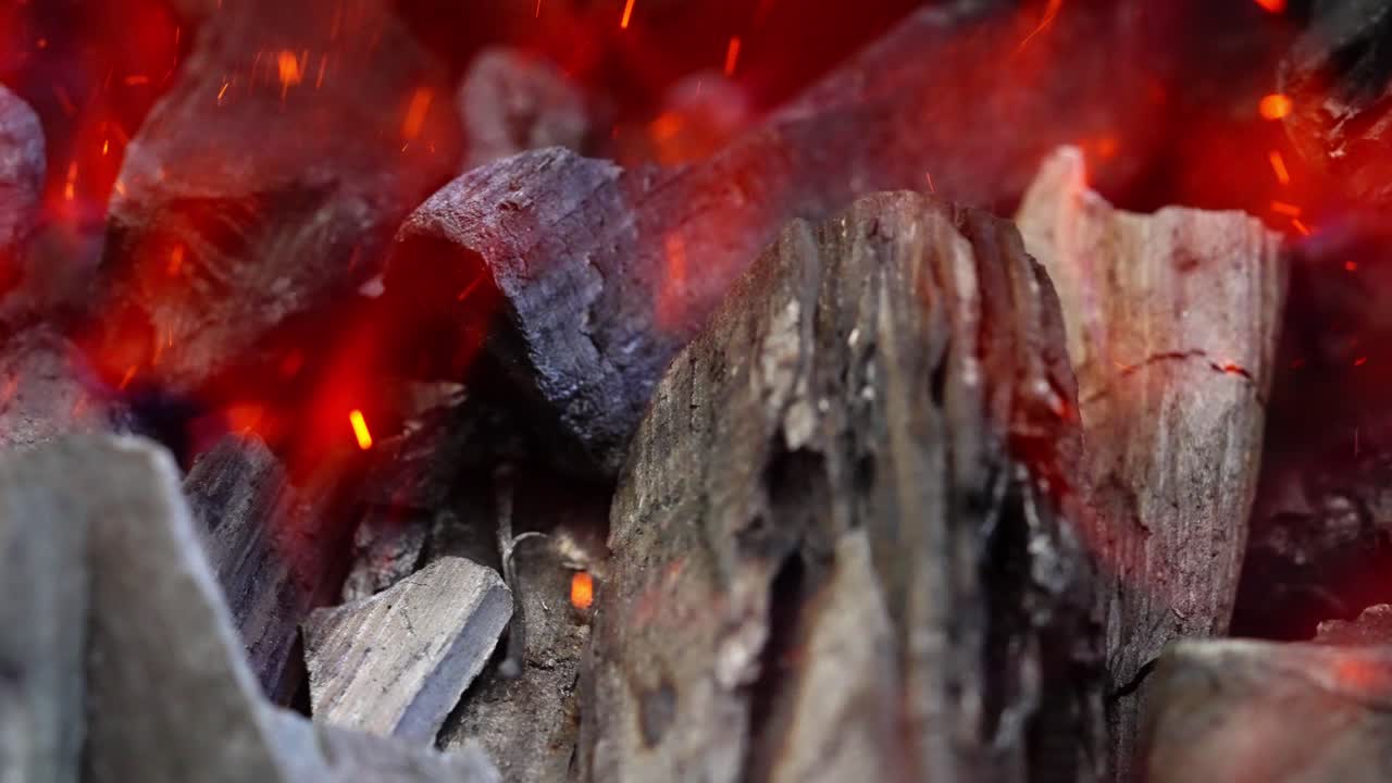 Close overview of glowing embers and rising flames in blackened grill pit, rustic cooking setting, slow motion closeup backdrop background