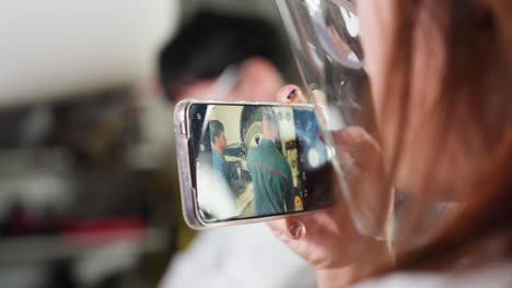 close-up of lady using phone to record mechanic working, seen through her smartphone screen with blurry view of colleague standing by in workshop