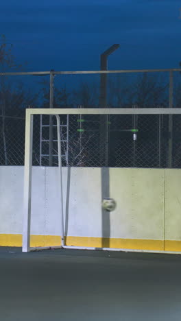 la pelota de fútbol entra en el poste de la portería con precisión y rueda en el campo al aire libre iluminado durante la práctica nocturna, rodeada por un paisaje urbano con altos edificios residenciales y luces vibrantes de la ciudad