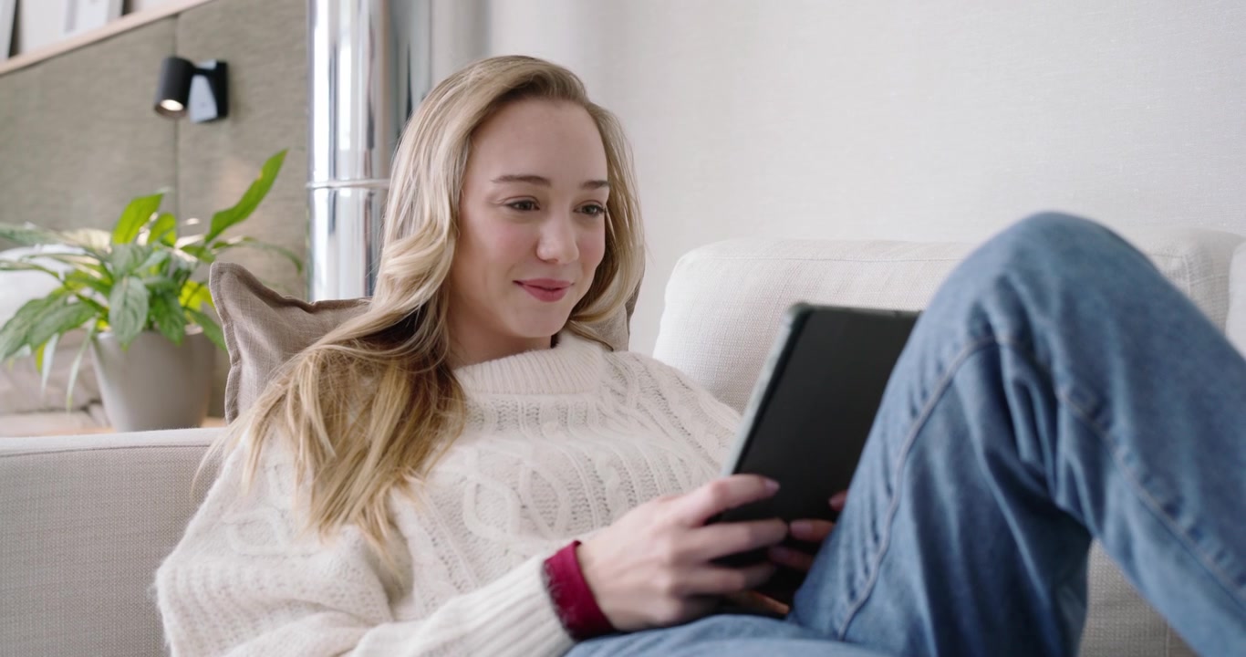 Kvinde i cremefarvet sweater og jeans, der ligger på sofaen, taster på tablet og kigger opmærksomt.