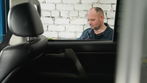 mechanic in blue uniform pressing lift button as car rises in garage, viewed through open car window, white brick wall and blue equipment visible in background