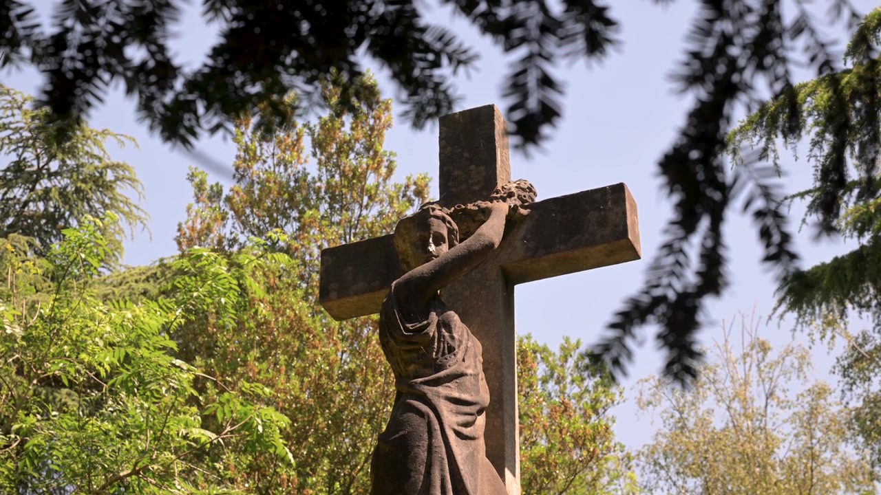 Un monumento in pietra con una croce è parzialmente incorniciato da alberi rigogliosi in un cimitero. Ideale per l'uso in documentari storici o religiosi