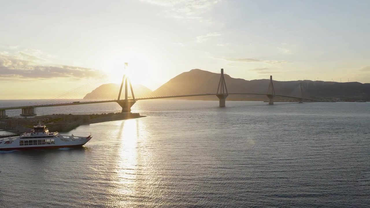 Ponte Rio-Antirrio sobre o Golfo de Patras ao pôr do sol perto de Patras, Grécia, brilho retroiluminado de drone em movimento acima de balsas de iates, crista da montanha.