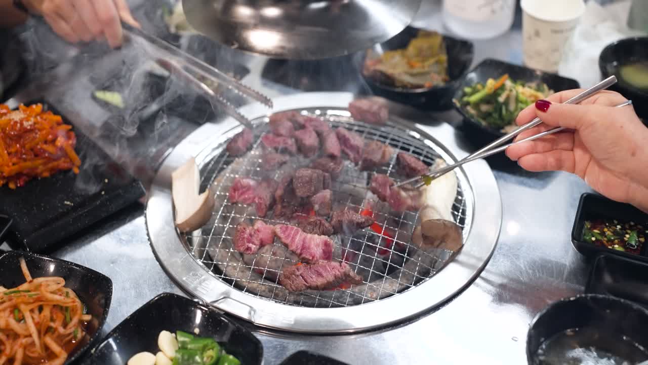 Close-up slow motion of high-quality Hanu beef being grilled on a sizzling charcoal grill surrounded by various delicious banchan side dishes in a vibrant Korean restaurant setting