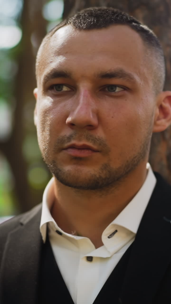 Man with short haircut and bald patches wearing classic black suit and white shirt with unbuttoned collar looks aside standing in city park closeup slow motion