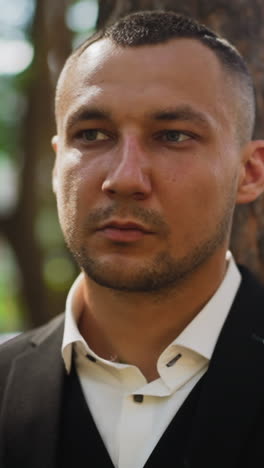 man with short haircut and bald patches wearing classic black suit and white shirt with unbuttoned collar looks aside standing in city park closeup slow motion