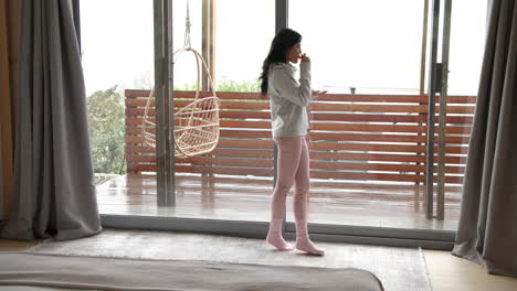 woman enjoying hot drink by window at home, wearing cozy winter clothes