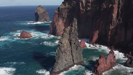 Rocks-and-cliffs-in-beautiful-blue-ocean-with-waves-crashing-in-creating-foam-during-sunny-day