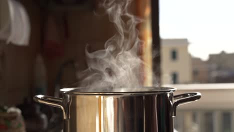 steaming and boiling pan of water on modern heating stove in kitchen on the background of open balcony. boiling with steam emitted from stainless cooking pot. evaporation effects in the air