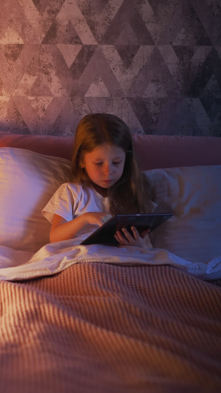 Pretty little child plays game online via tablet in comfortable bed with pillows and warm bedcover near illuminated play house in shaded room slow motion