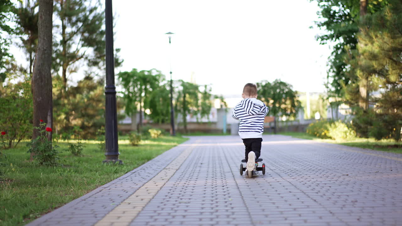 Caucasian male kid distancing from camera riding his scooter. Toddler spends time actively during the walk. Low angle view. Vertical video.