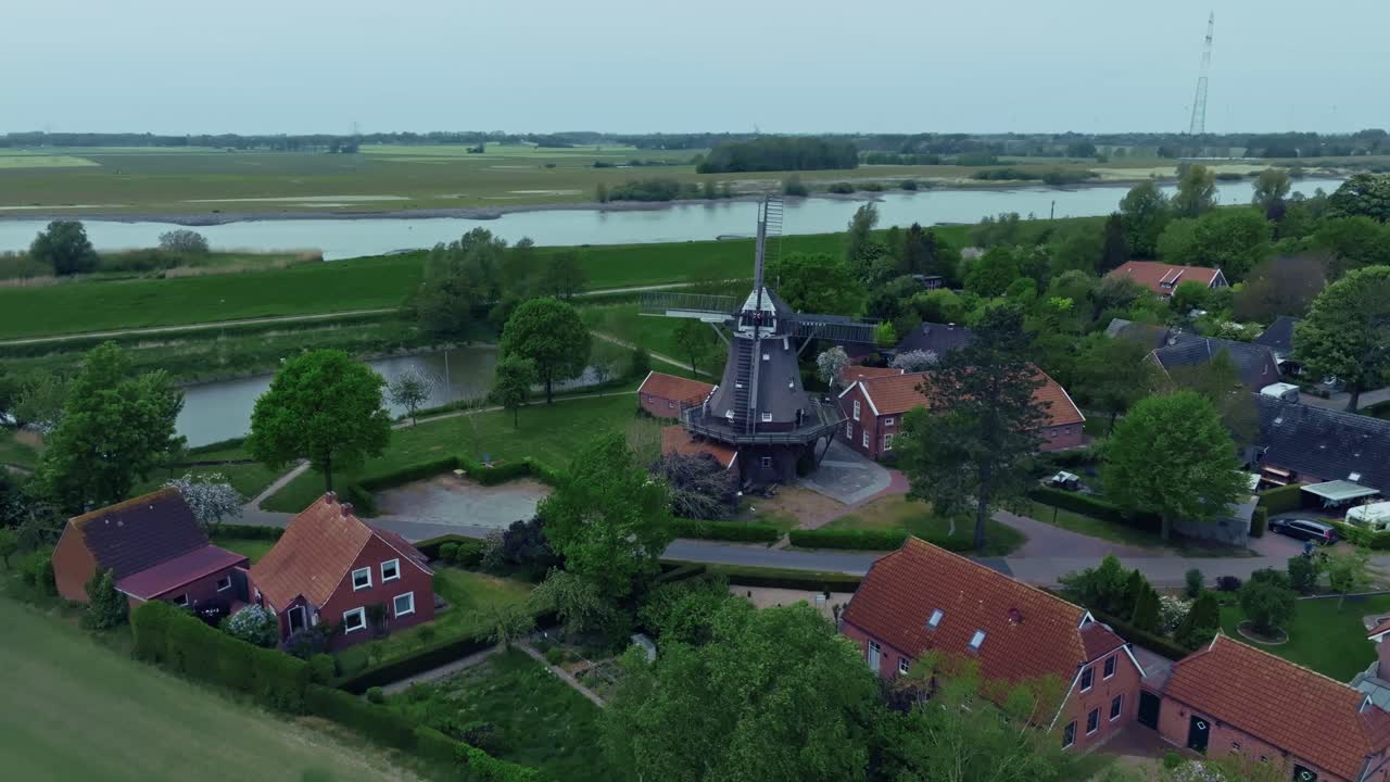 Drone pans right showing extensive green fields, a river and red-roof houses surrounding a windmill. Bright daylight with slight haze.