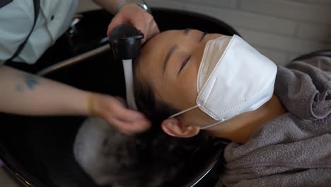 Friseur-Wäscht-Schaum-Von-Den-Haaren-Einer-Asiatischen-Frau,-Die-Im-Schönheitssalon-Eine-Virenschützende-Gesichtsmaske-Trägt