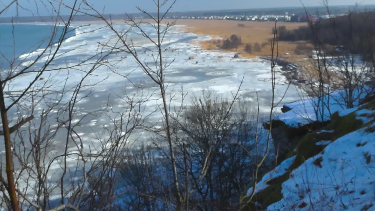 Icy and frozen brown grassy beach shoreline in Tiskre with chunks and pieces of ice in Baltic blue ocean sea water during a sunny day viewed from high altitude Tabasalu bank cliff coast through trees.