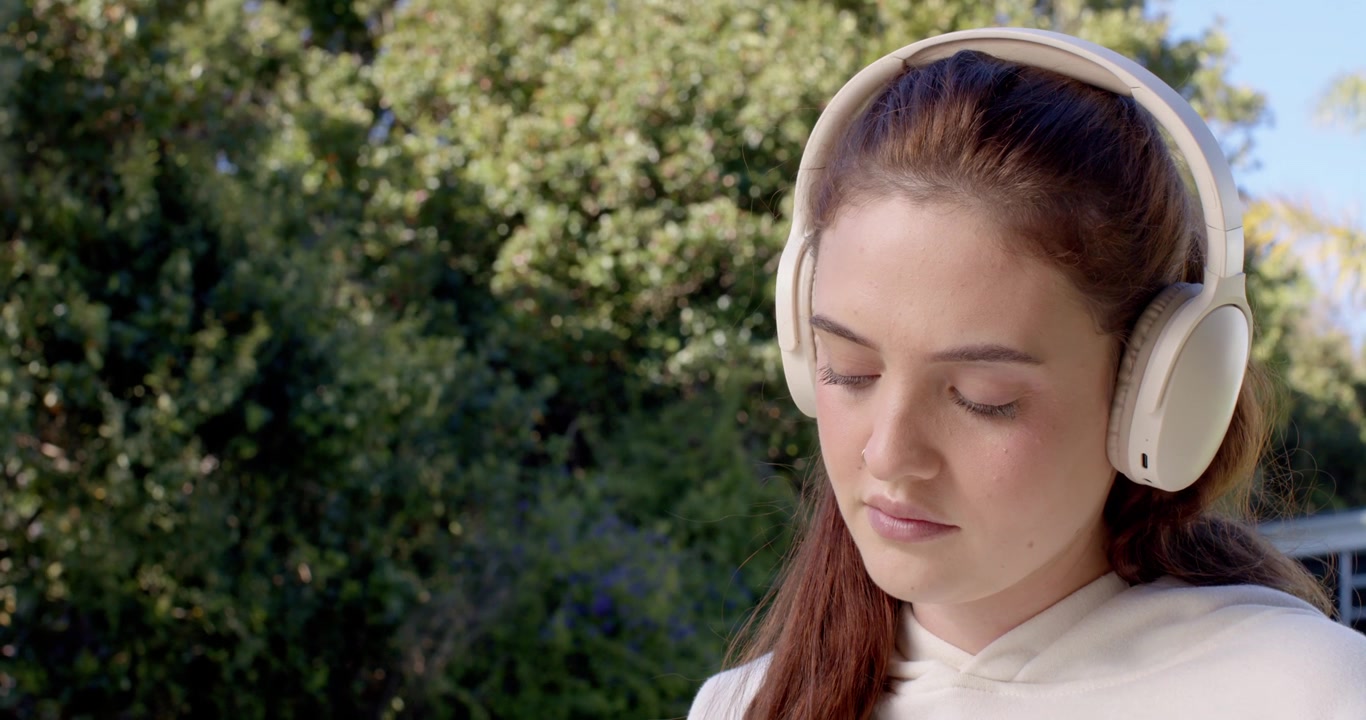 Donna che indossa una felpa leggera con cappuccio e cuffie over-ear bianche, con lo sguardo rivolto verso il basso sul portico.